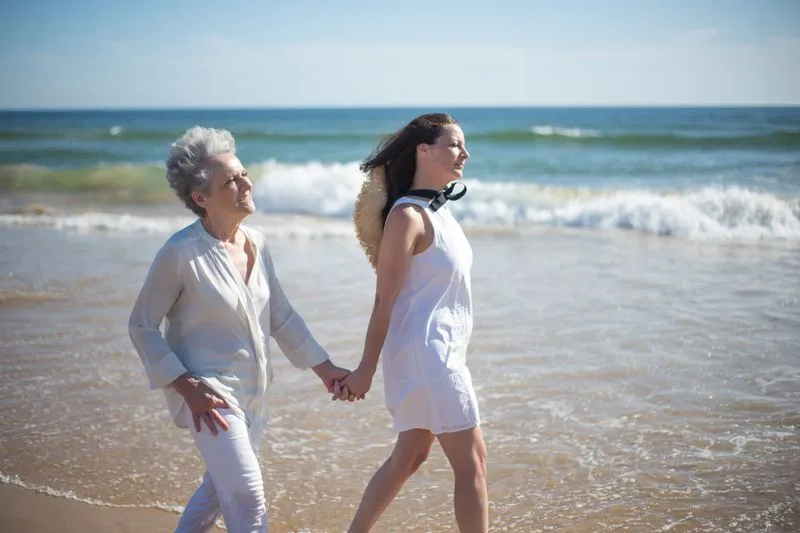 Femme brune et femme âgée aux cheveux blancs qui marchent main dans la main au bord de l'eau, sur une plage ensoleillée