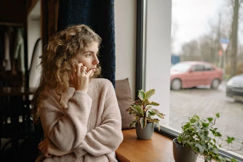 Femme assise sur une banquette près d'une fenêtre, le téléphone à l'oreille, le regard tourné vers l'extérieur