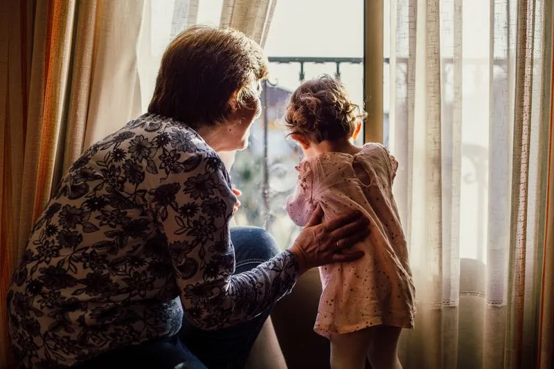 Femme âgée vue de dos qui retient d'une main une petite fille debout devant une fenêtre aux voilages clairs