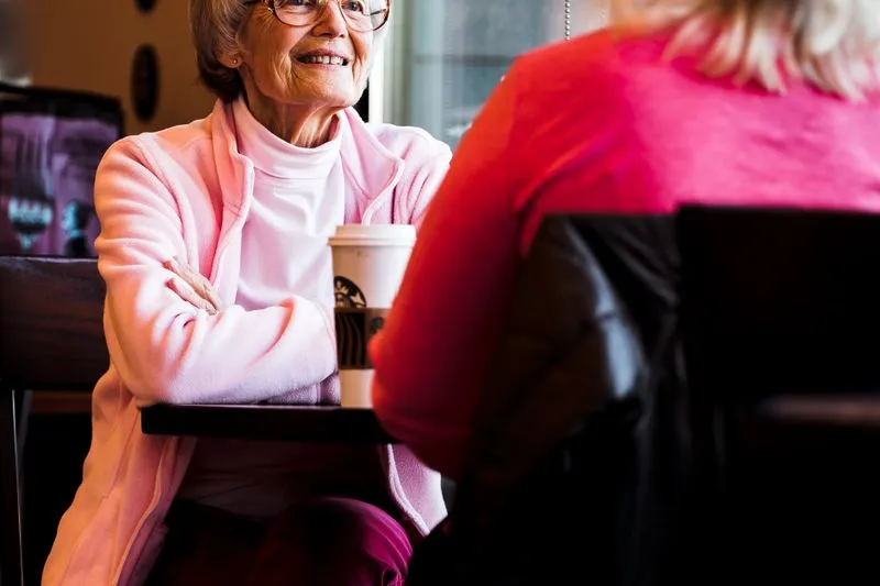 Femme âgée souriante attablée dans un café, face à une femme vue de dos, un gobelet à emporter posé sur la table