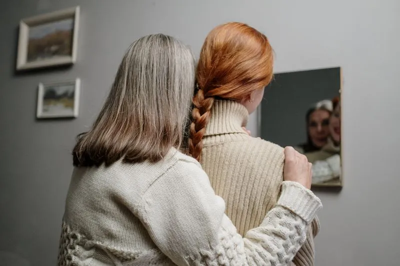Femme aux cheveux gris, la main posée sur l'épaule de sa fille adulte à la tresse rousse, toutes deux de dos face à un miroir