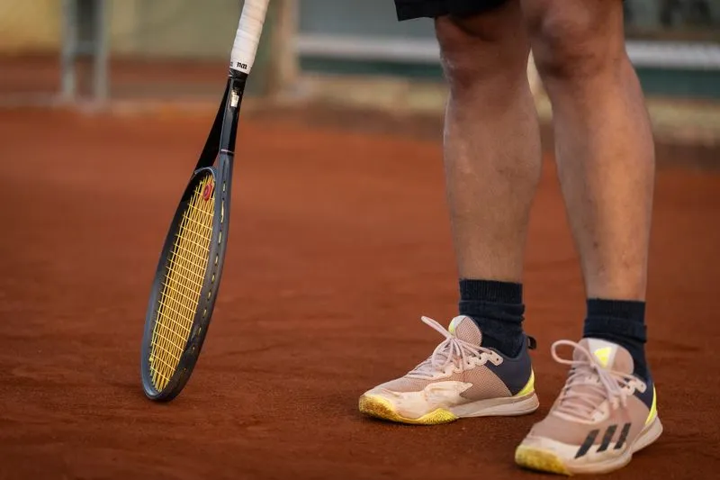 Gros plan sur les jambes d'un joueur de tennis, baskets couvertes de terre battue, raquette posée sur le court