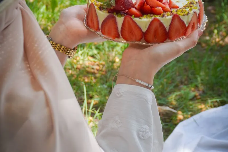 Fraisier entier tenu à deux mains dans un jardin, moitiés de fraises plaquées sur tout le pourtour, dessus garni de fraises et de pistaches concassées