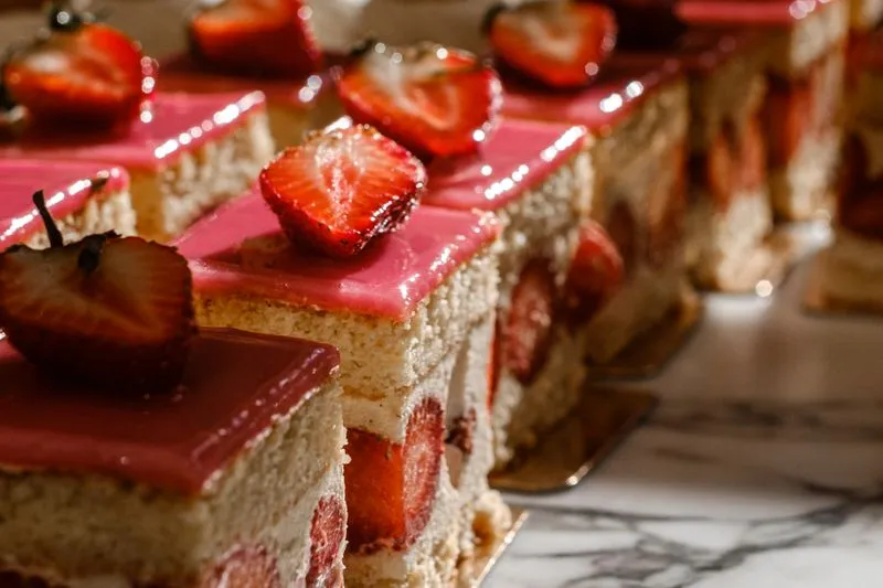 Parts carrées de fraisier alignées sur un plan en marbre : génoise, crème et fraises coupées visibles sur la tranche, glaçage rose et demi-fraise sur le dessus