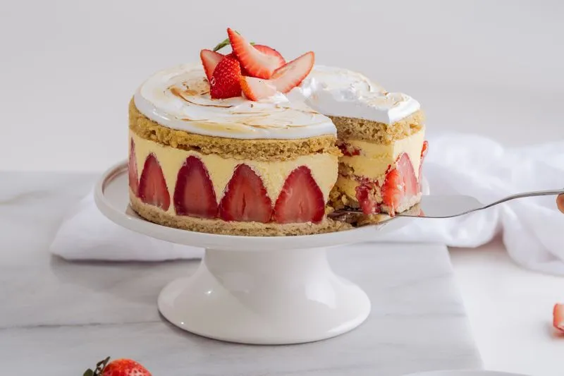 Fraisier sur un présentoir blanc : moitiés de fraises alignées sur le pourtour, dessus de meringue dorée garni de fraises, une part retirée à la pelle à tarte