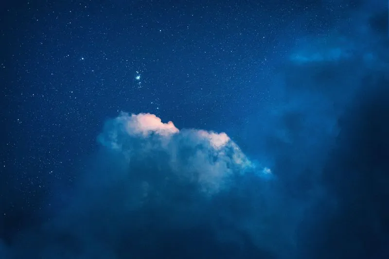 Ciel nocturne bleu constellé d'étoiles, avec une petite nébuleuse lumineuse au-dessus d'un nuage éclairé par le crépuscule