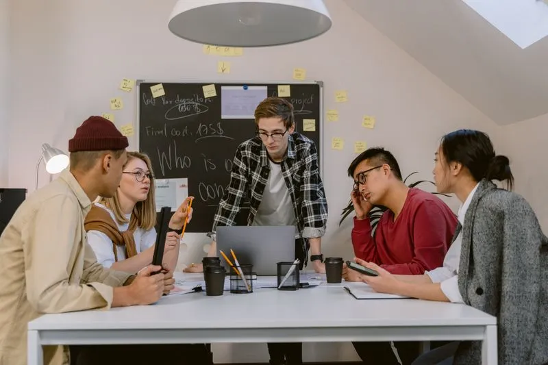 Jeune homme qui présente un projet devant un tableau couvert de post-it, entouré de collègues assis autour d'une table