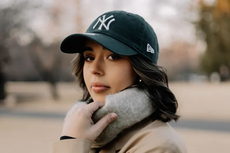 Jeune femme en casquette en coton vert foncé brodée, écharpe en laine grise et trench beige, dans un parc