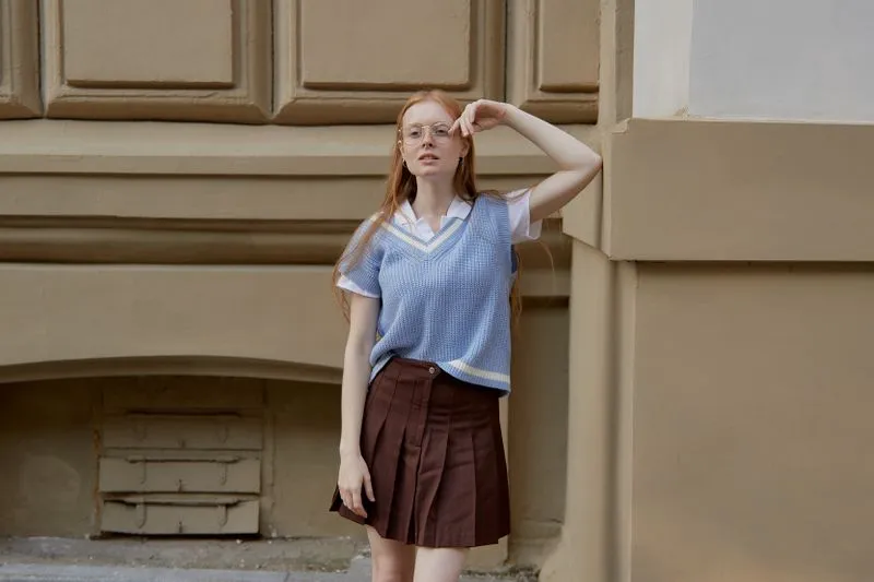 Jeune femme en jupe plissée brune et pull sans manches à col V bleu clair porté sur une chemise blanche