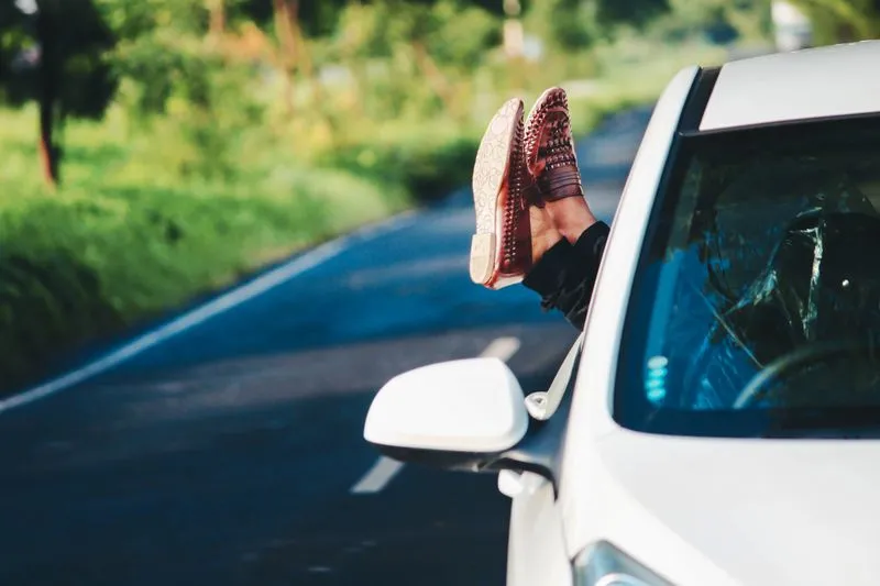 Pieds chaussés de mocassins en cuir tressé, sortis par la vitre d'une voiture sur une route de campagne