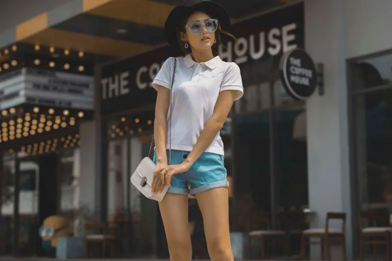 Jeune femme en polo blanc et short en jean, chapeau noir et lunettes de soleil, devant la façade d'un café