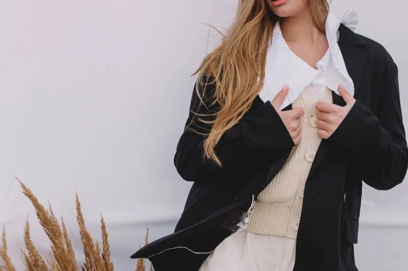 Blazer sombre ouvert sur une chemise blanche à col volanté, un gilet en maille écru boutonné et un pantalon crème
