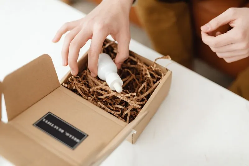 Main qui sort d'un colis en carton un petit flacon compte-gouttes blanc, sans étiquette