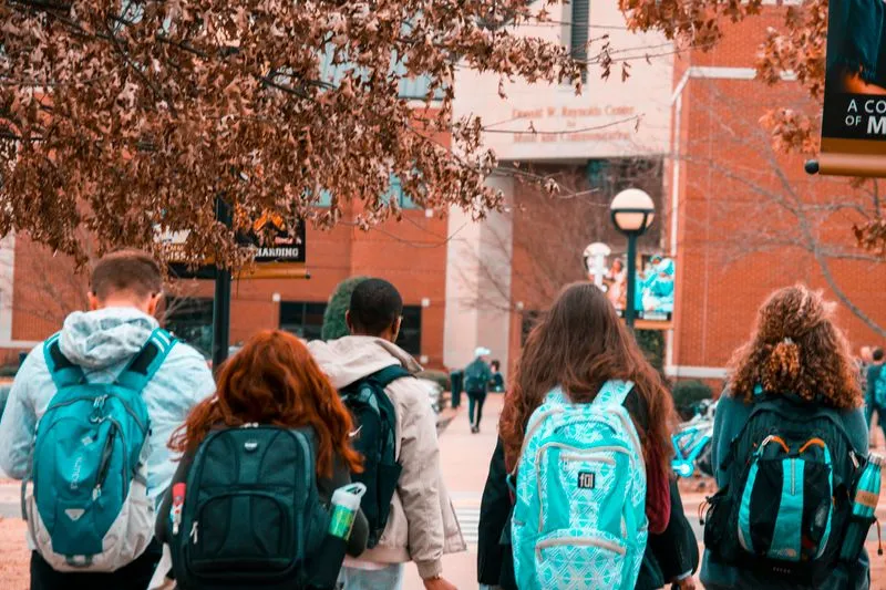 Groupe de jeunes vus de dos, sac à dos sur les épaules, marchant vers un bâtiment scolaire en brique
