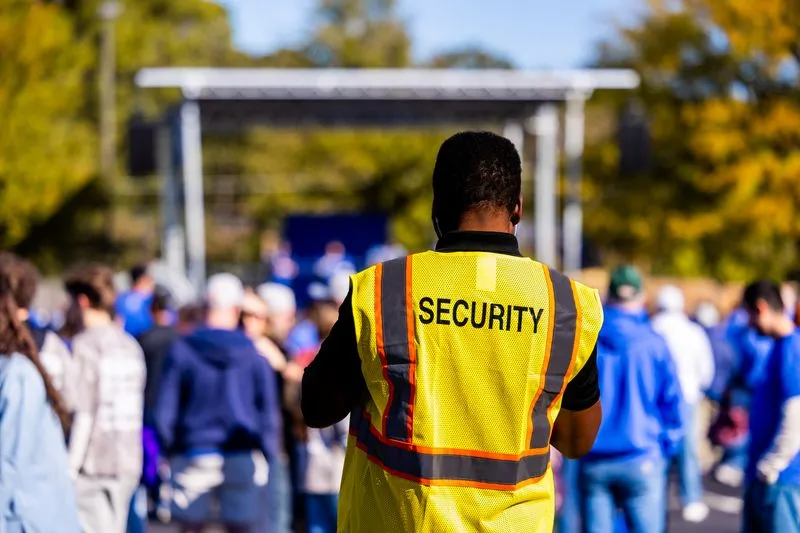 Agent de sécurité vu de dos, gilet jaune floqué « Security », au milieu du public d'un événement en plein air, en plein jour, devant une structure de scène vide