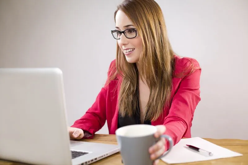 Jeune femme souriante assise devant un ordinateur portable, une tasse de café à la main près du clavier