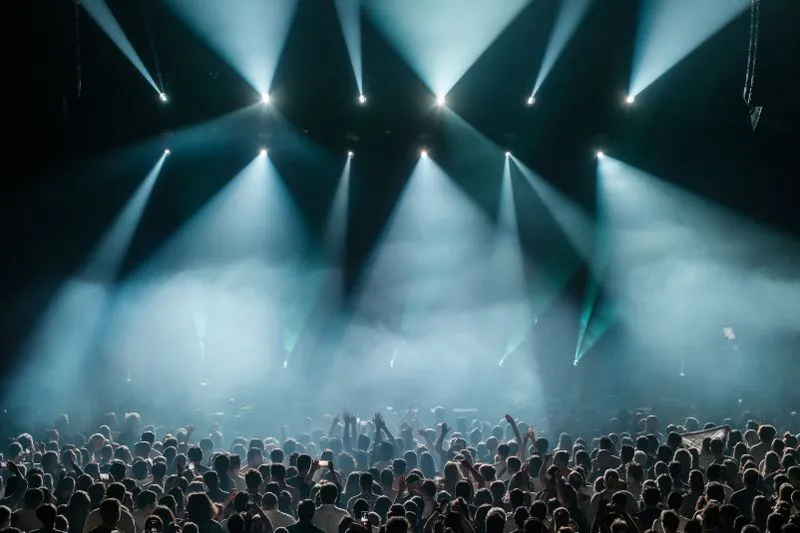 Foule compacte lors d'un concert, bras levés vers une scène traversée de faisceaux de lumière bleutée et de fumée