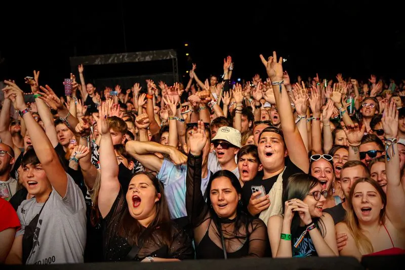 Foule dense à un concert de nuit, vue depuis la scène, bras levés et visages tournés vers les artistes, quelques téléphones brandis