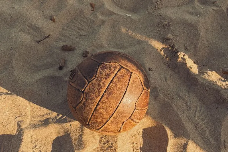 Vieux ballon de football en cuir brun, usé, à longs panneaux cousus, posé dans le sable