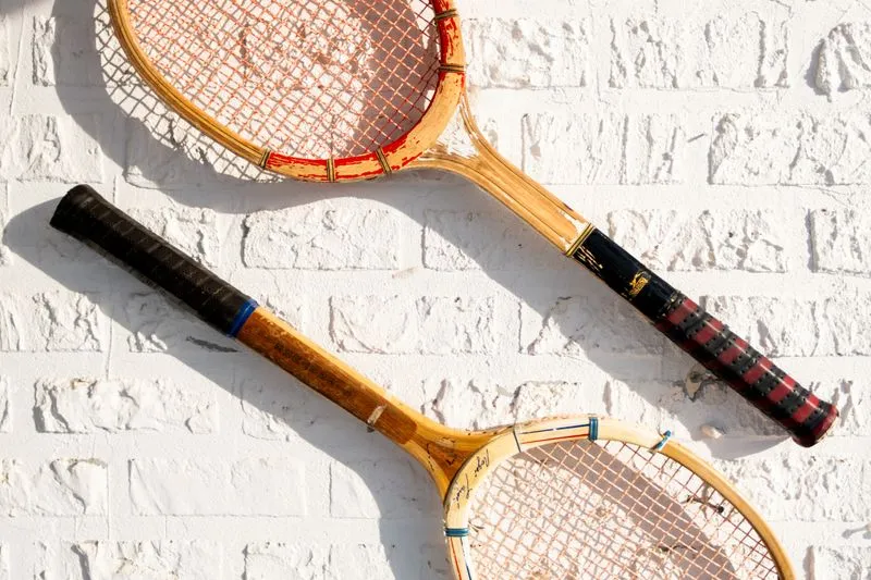 Deux raquettes de tennis anciennes en bois, aux manches gainés, accrochées au soleil sur un mur de briques peint en blanc