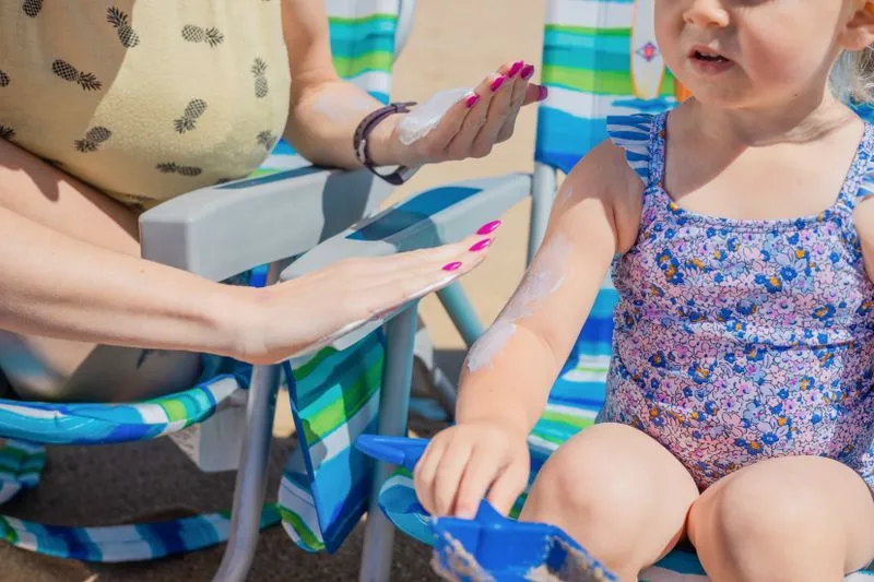 Une adulte étale une crème solaire blanche et épaisse sur le bras d'une fillette, à la plage