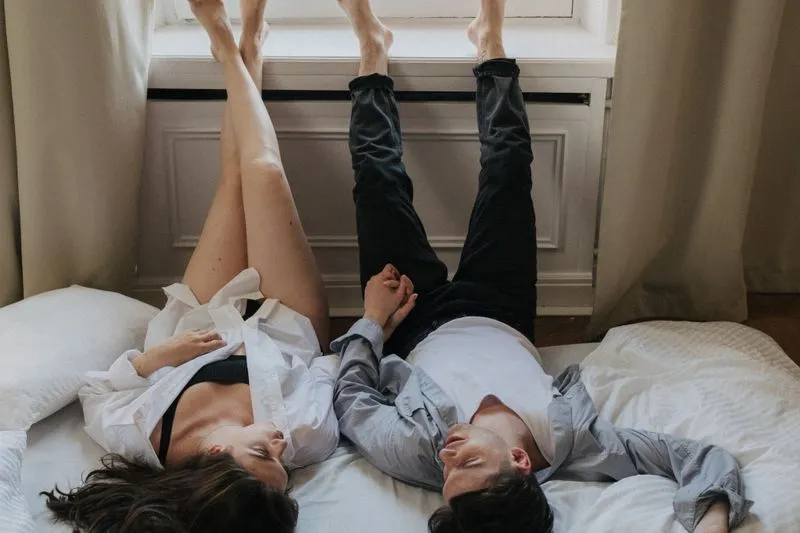 Couple allongé sur le dos sur un lit, jambes levées contre le rebord d'une fenêtre, main dans la main