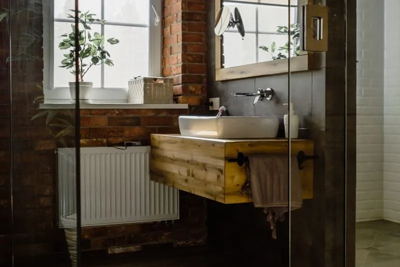 Plante verte en pot sur le rebord de la fenêtre d'une salle de bains, à côté d'une vasque posée sur un meuble en bois et d'un mur de briques