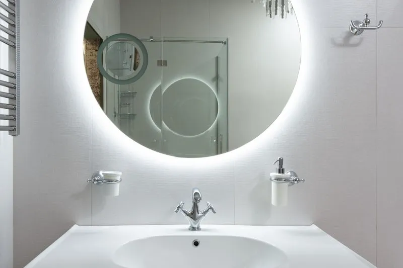 Lavabo blanc sous un grand miroir rond rétroéclairé, dans lequel se reflète une douche vitrée
