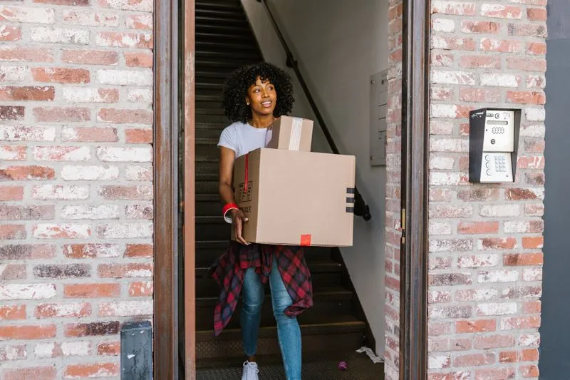 Jeune femme souriante qui sort d'un immeuble en portant un carton, prête pour un nouveau départ
