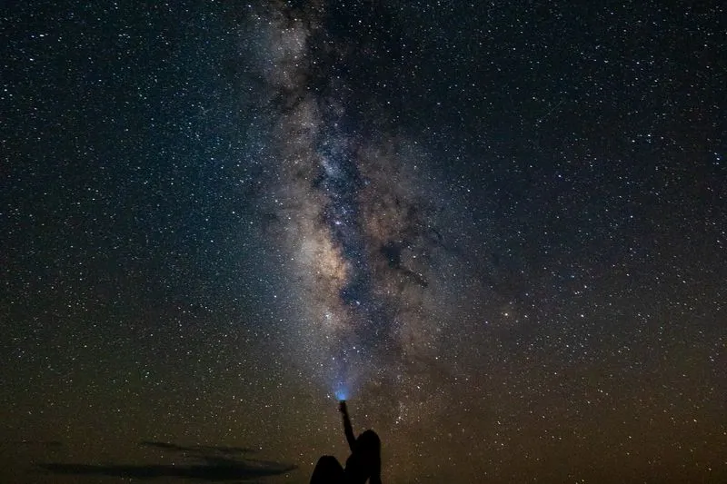 Silhouette d'une personne assise dehors la nuit, bras levé vers un ciel étoilé traversé par la Voie lactée