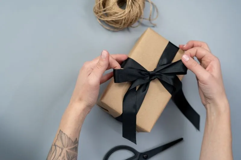 Mains qui nouent un ruban noir autour d'un petit paquet emballé de papier kraft, une pelote de ficelle posée à côté