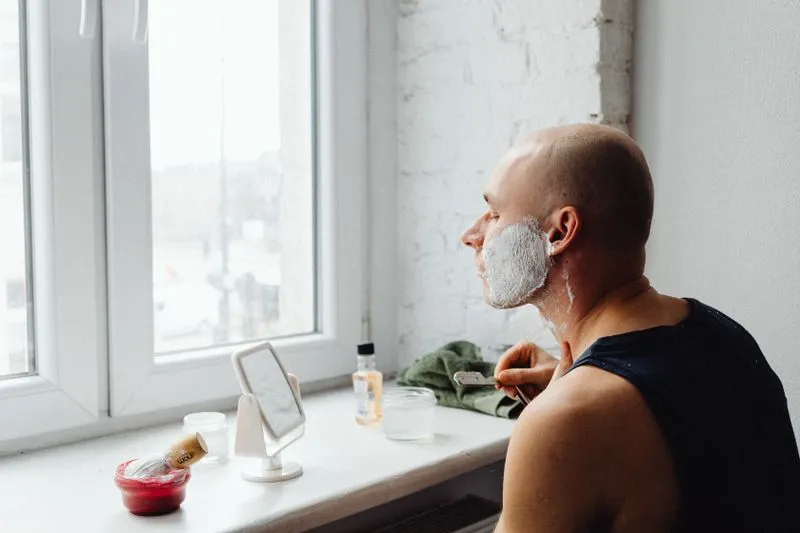 Homme en débardeur qui se rase près d'une fenêtre, mousse à raser sur le visage, face à un petit miroir posé sur le rebord