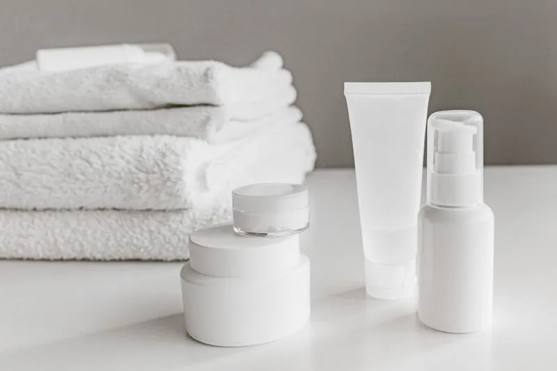 Pots de crème empilés, un tube et un flacon pompe blancs posés sur un plan de toilette, des serviettes pliées en arrière-plan