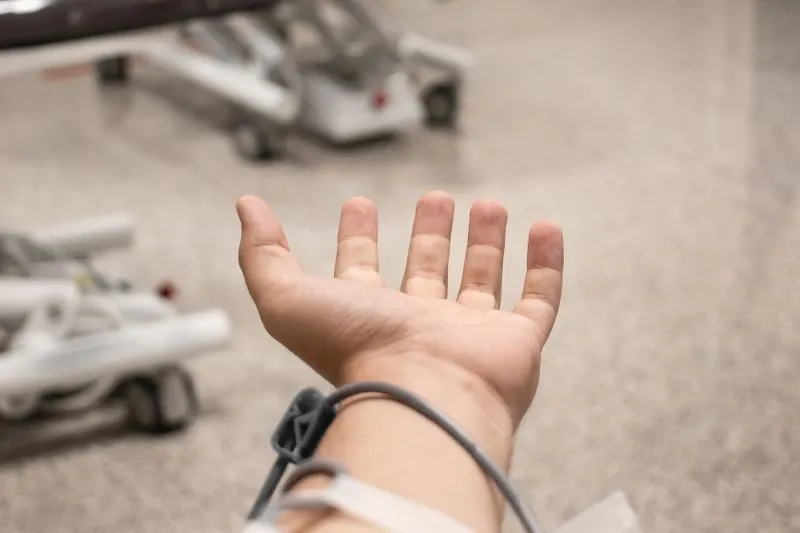 Main ouverte et avant-bras, un tube gris enroulé autour du poignet, devant du matériel médical sur roulettes