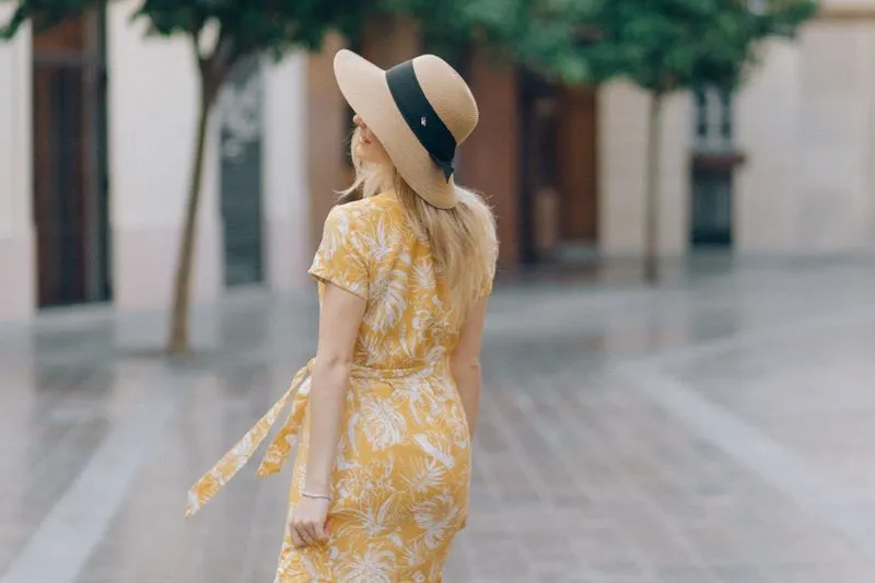 Femme de dos en robe jaune imprimée et chapeau de paille à ruban noir, sur une place pavée