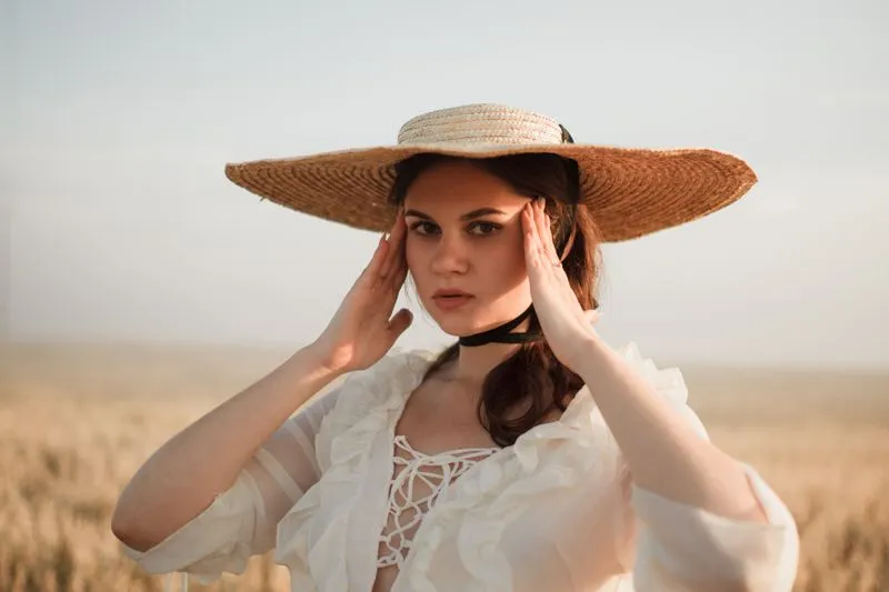 Femme en robe blanche à volants et chapeau de paille à large bord, mains sur les tempes, dans un champ de blé sous le soleil