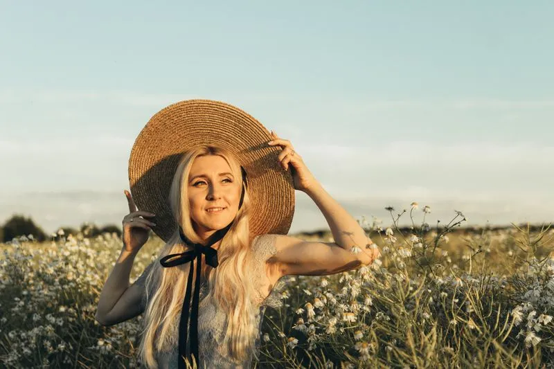 Femme souriante en chapeau de paille à large bord et ruban noir, tenant le bord du chapeau, dans un champ de fleurs sauvages