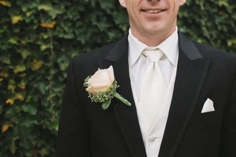 Gros plan sur un costume noir, gilet et cravate crème, boutonnière d'une rose pâle, devant un mur de lierre