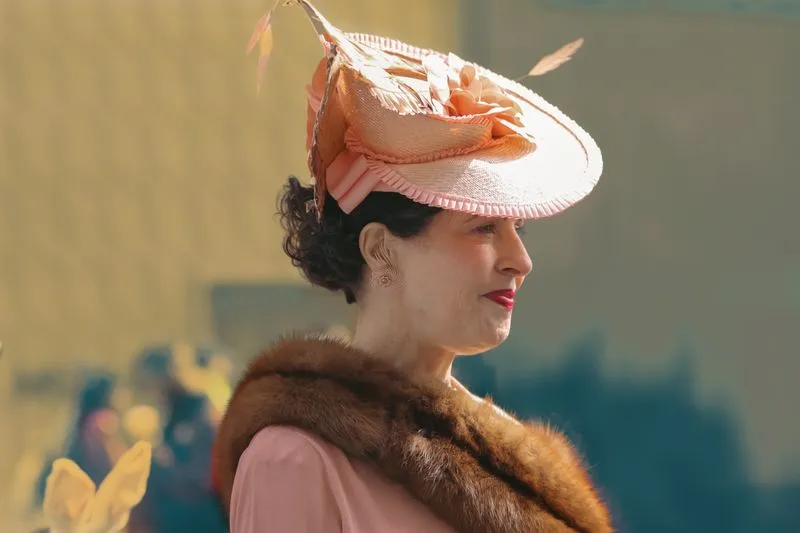 Femme de profil en tenue rose, grand chapeau de paille rose orné de plumes et étole de fourrure sur les épaules