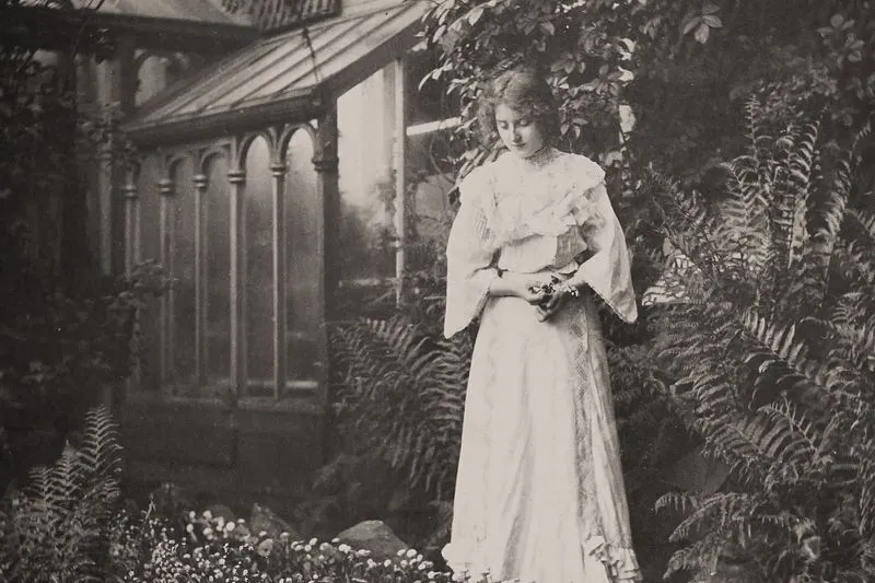 Photographie ancienne sépia d'une femme en longue robe blanche à volants et manches évasées, debout parmi les fougères devant une serre