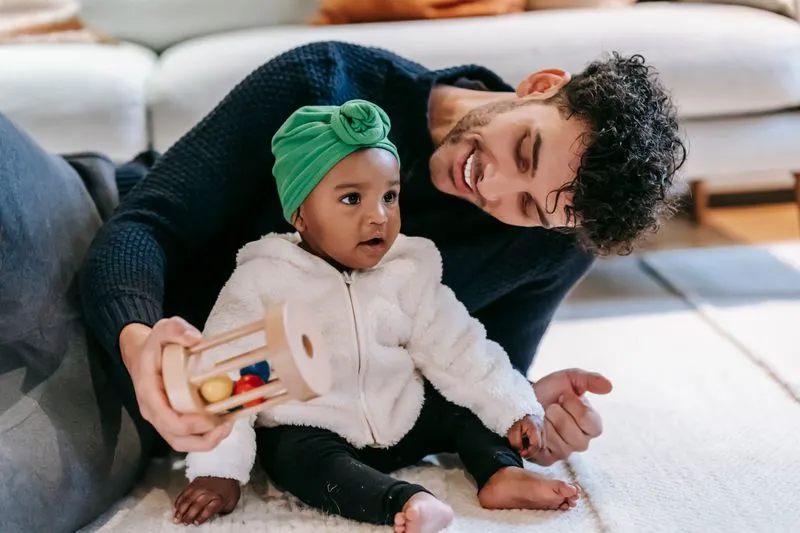 Père allongé au sol, tenant un jouet, souriant à son bébé assis sur un tapis