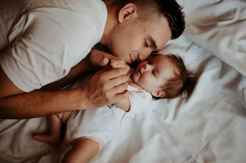Jeune père allongé sur un lit, les yeux fermés, le visage contre celui de son bébé