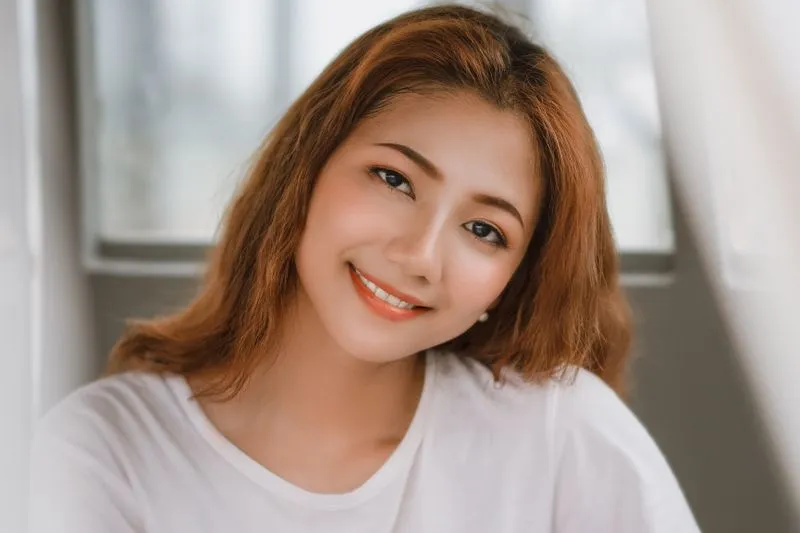 Portrait d'une jeune femme souriante en tee-shirt blanc uni, devant une fenêtre lumineuse