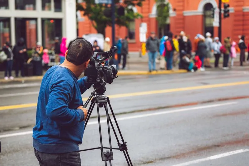 Un cadreur filme une rue avec une caméra sur trépied, face à des passants en imperméable massés sur le trottoir d'en face