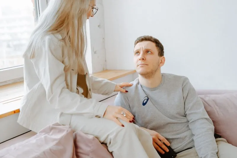 Une femme debout près d'un canapé pose la main sur l'épaule de son compagnon assis, qui lève vers elle un regard inquiet
