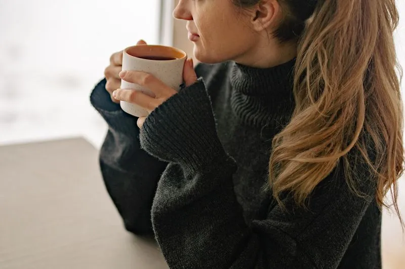 Une femme en pull sombre à col roulé, une tasse entre les mains, tournée vers une fenêtre