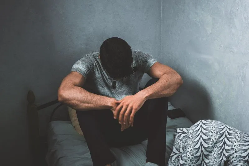 Un homme assis au bord d'un lit, les coudes sur les genoux, le regard baissé dans une chambre en pénombre