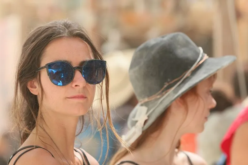 Deux jeunes femmes dans une foule de festival en plein soleil, l'une en lunettes de soleil, l'autre en chapeau à large bord