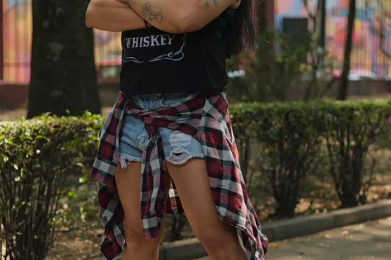 Jeune femme en débardeur noir et short en jean troué, bras croisés, une chemise à carreaux rouge et noir nouée autour de la taille