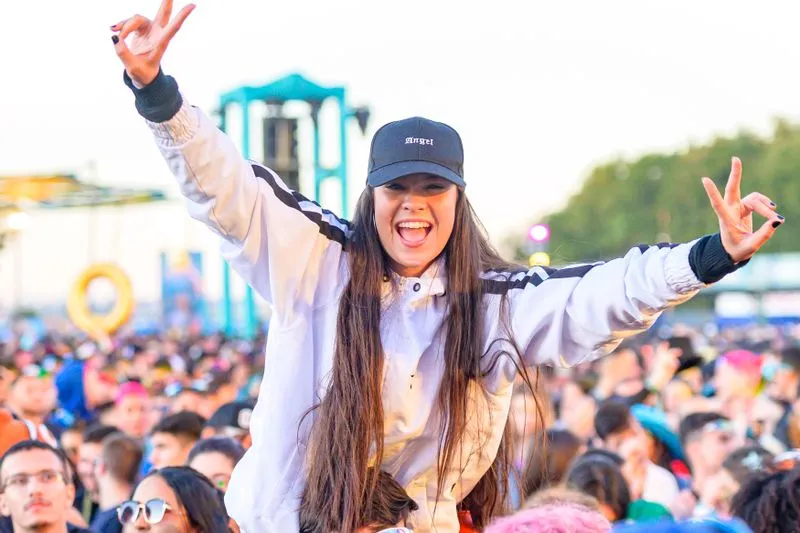 Jeune femme juchée sur les épaules d'un ami, casquette vissée sur la tête et bras levés en signe de paix, au milieu d'une foule de festival en fin de journée